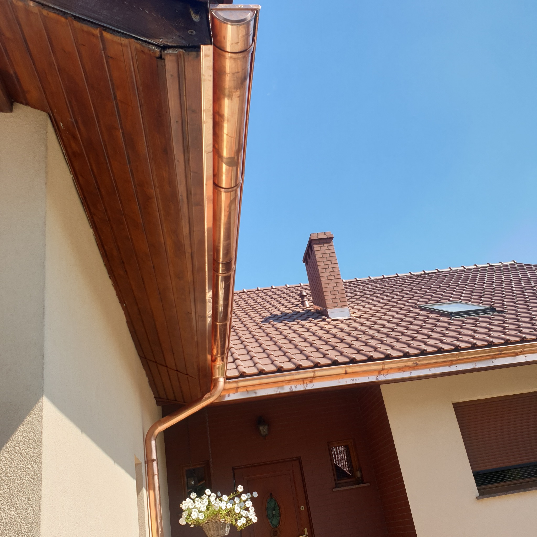 Miedziana rynna z pionowym odpływem na tle błękitnego nieba, widok z dołu na fragment dachu z brązową dachówką i kominem z cegły klinkierowej.