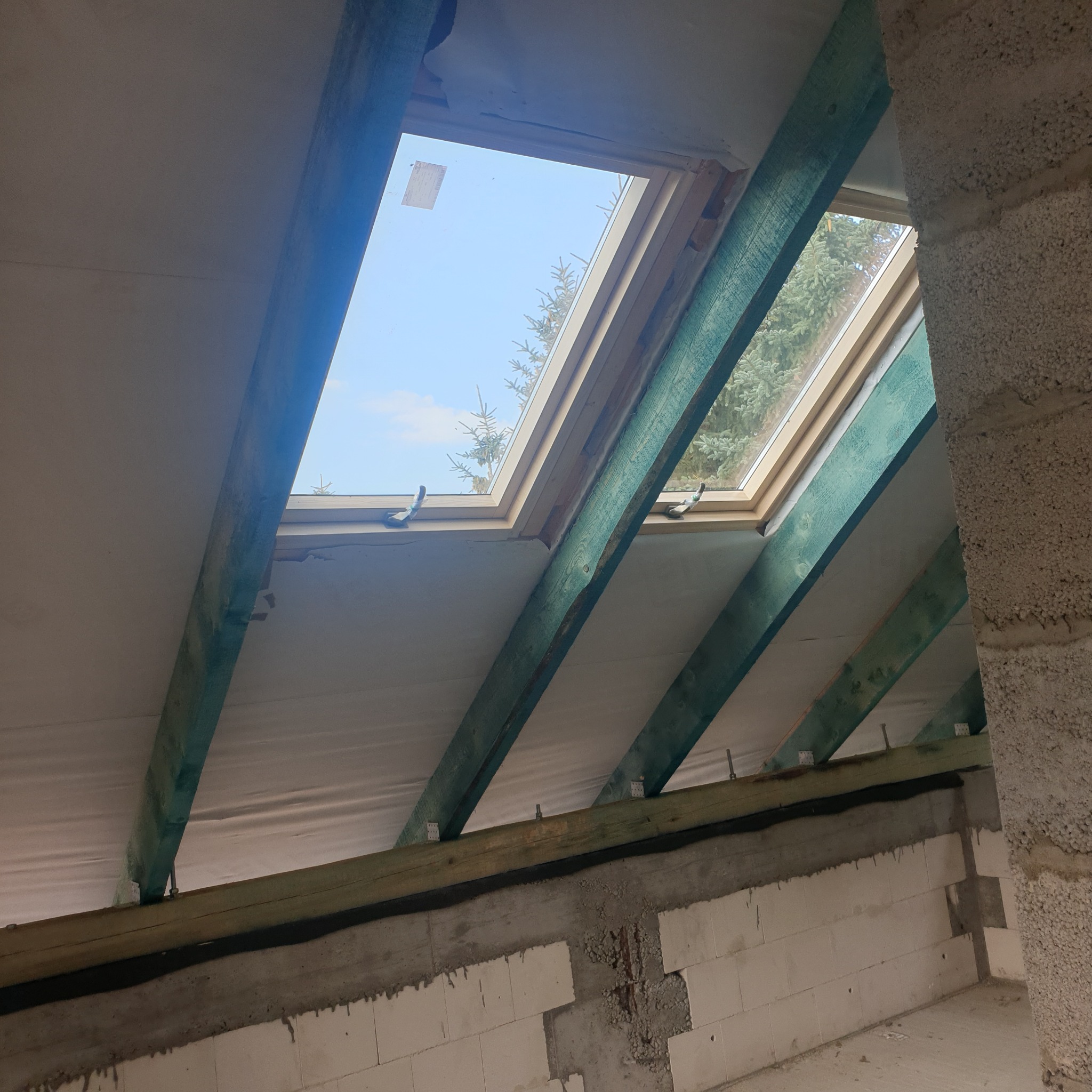 Poddasze w trakcie adaptacji z widocznymi krokwiami pomalowanymi na zielono, oknami dachowymi z widokiem na drzewa i niebo oraz ścianami z betonu komórkowego.