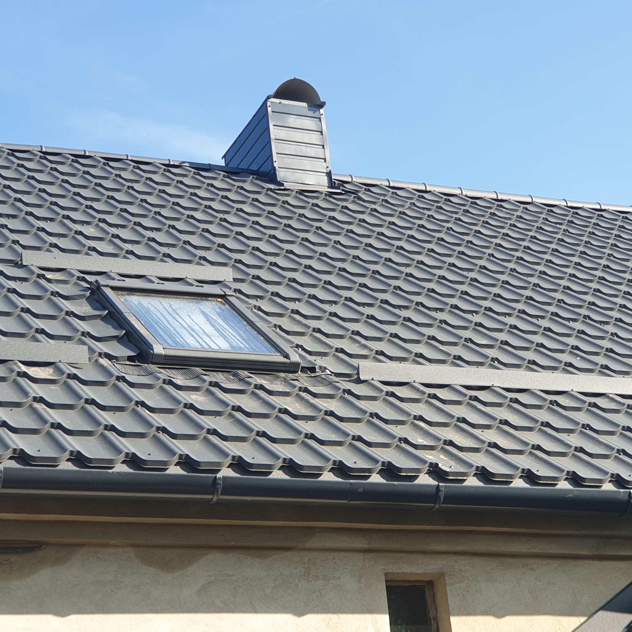 Grafitowy dach z blachodachówki z oknem dachowym i kominem wentylacyjnym, widok z dołu na tle błękitnego nieba.