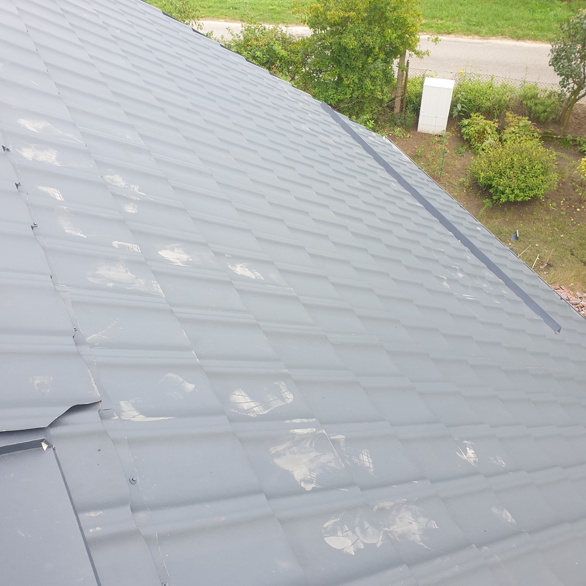 Szary dach z blachy trapezowej widziany z góry, z widocznymi elementami obróbki blacharskiej przy krawędzi i refleksami światła na powierzchni, w tle zieleń ogrodu.
