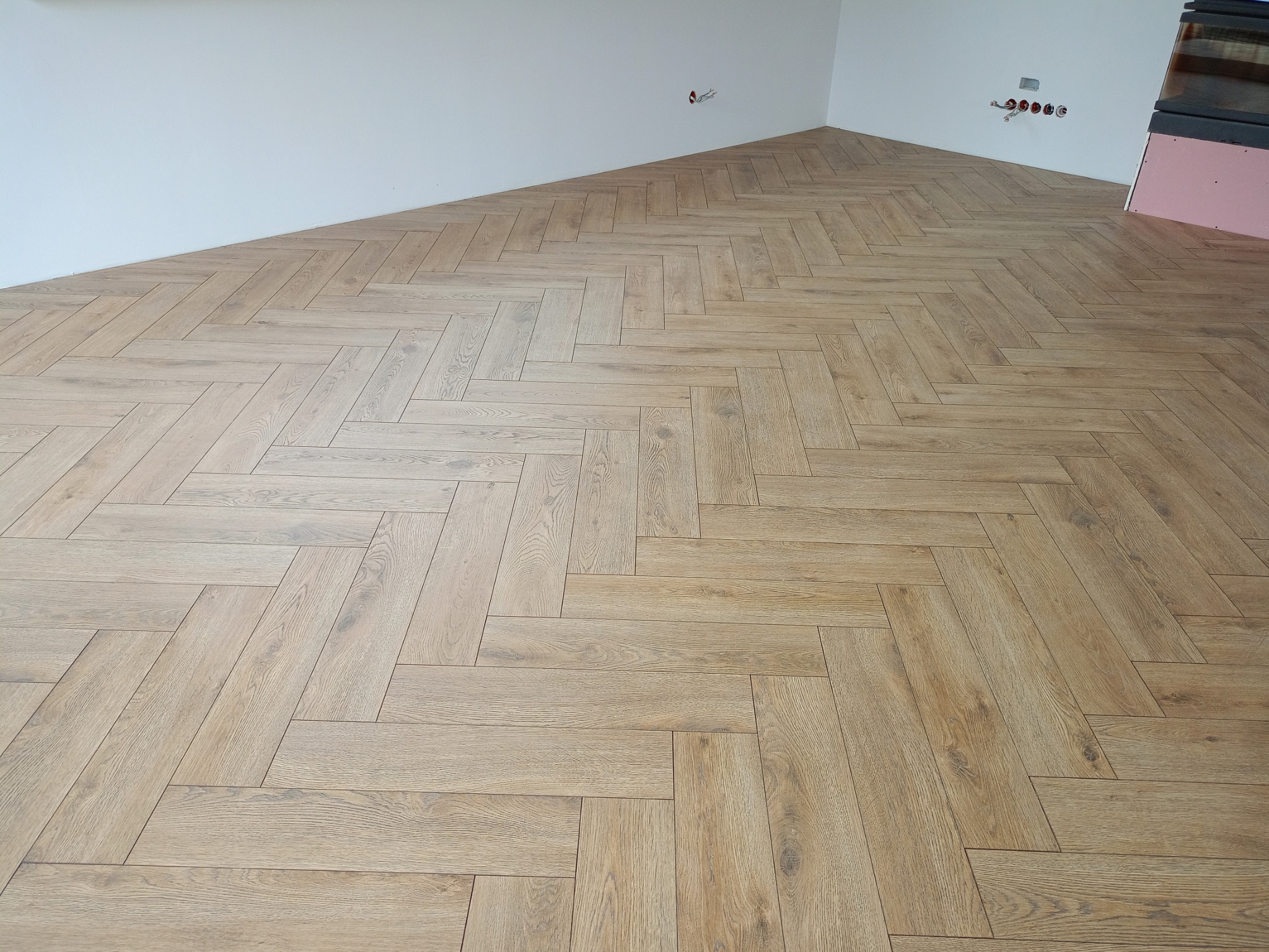 Ułożona podłoga z paneli w jodełkę w jasnym odcieniu drewna, widoczne świeżo pomalowane, białe ściany i wystające kable elektryczne.