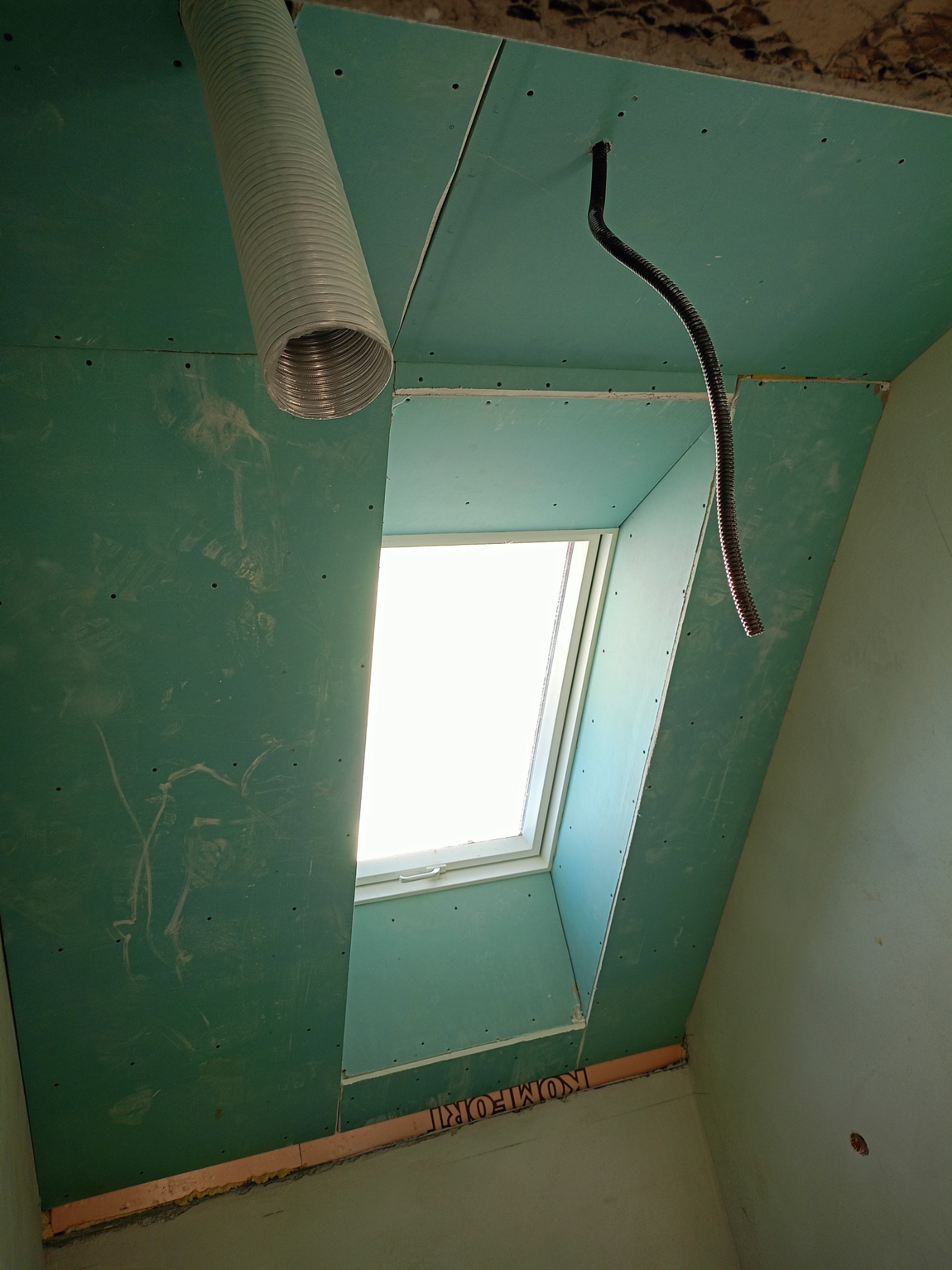 Poddasze z oknem dachowym w trakcie prac wykończeniowych: płyty kartonowo-gipsowe zamontowane na skosach, widoczny przewód wentylacyjny i instalacja elektryczna.