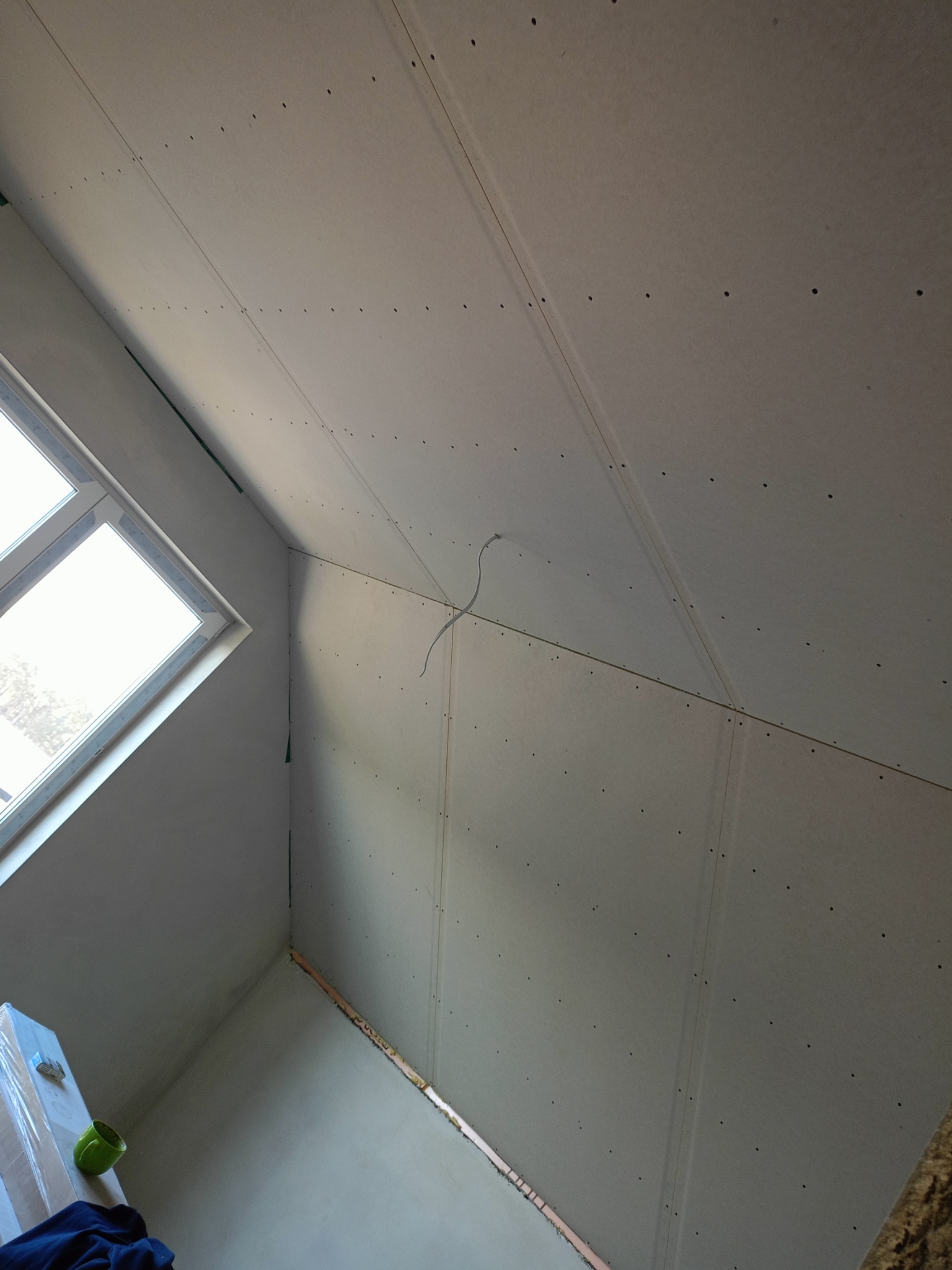 Skośny sufit poddasza z zamontowaną płytą kartonowo-gipsową, widoczne łączenia płyt i ślady po wkrętach, okno dachowe w lewym dolnym rogu, fragment drabiny i kubek na pierwszym planie.