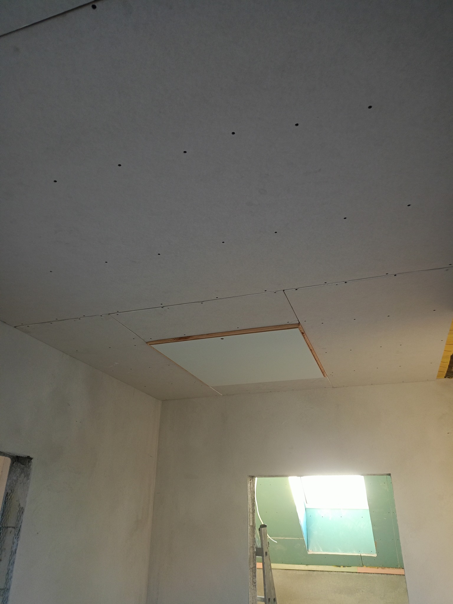 Sufit podwieszany z płyty karton gips, widoczne wkręty, otwór rewizyjny z drewnianą ramą, ściany surowe, widok na kolejne pomieszczenie z drabiną.
