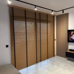 Dawid Kurek - Zamknięte drewniane żaluzje z czarnymi sznurkami w nowoczesnym salonie z drewnianą ścianą dekoracyjną i oświetleniem szynowym.