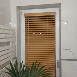 Dawid Kurek - Jasnobrązowe listewki w oknie, z białymi ramami i marmurowymi płytkami w tle, oraz zielona roślina w dolnym rogu.