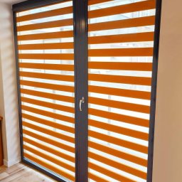 Dawid Kurek - Pomarańczowe rolety w systemie dzień i noc, zamontowane w dwuskrzydłowym oknie z ciemną ramą, widoczne z perspektywy wnętrza pomieszczenia.