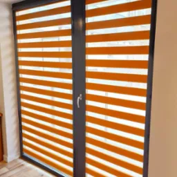 Pomarańczowe rolety w systemie dzień i noc, zamontowane w dwuskrzydłowym oknie z ciemną ramą, widoczne z perspektywy wnętrza pomieszczenia.