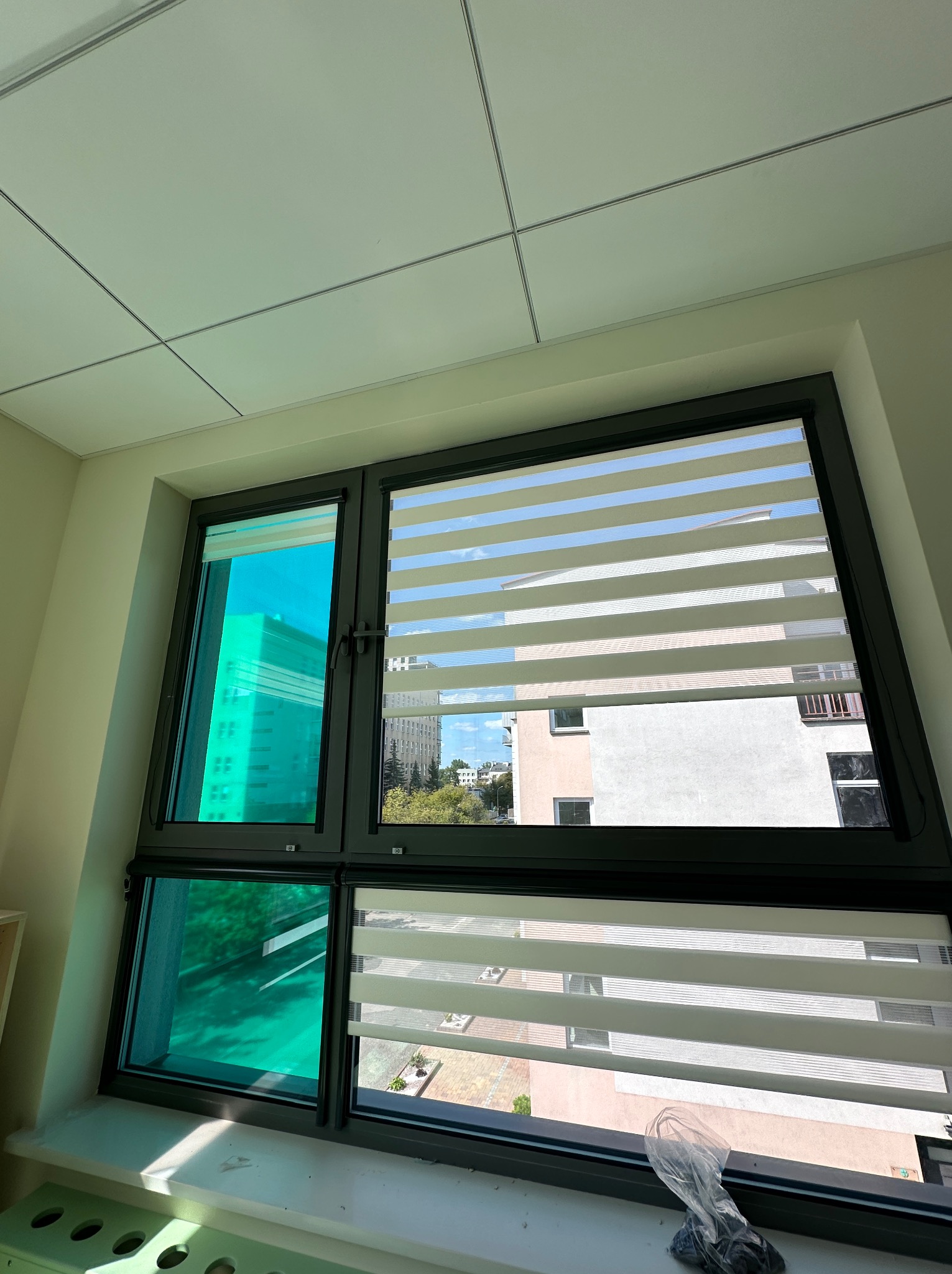 Okno z zamontowanymi roletami dzień noc, jedna część okna z folią okienną, widok na budynki na zewnątrz.