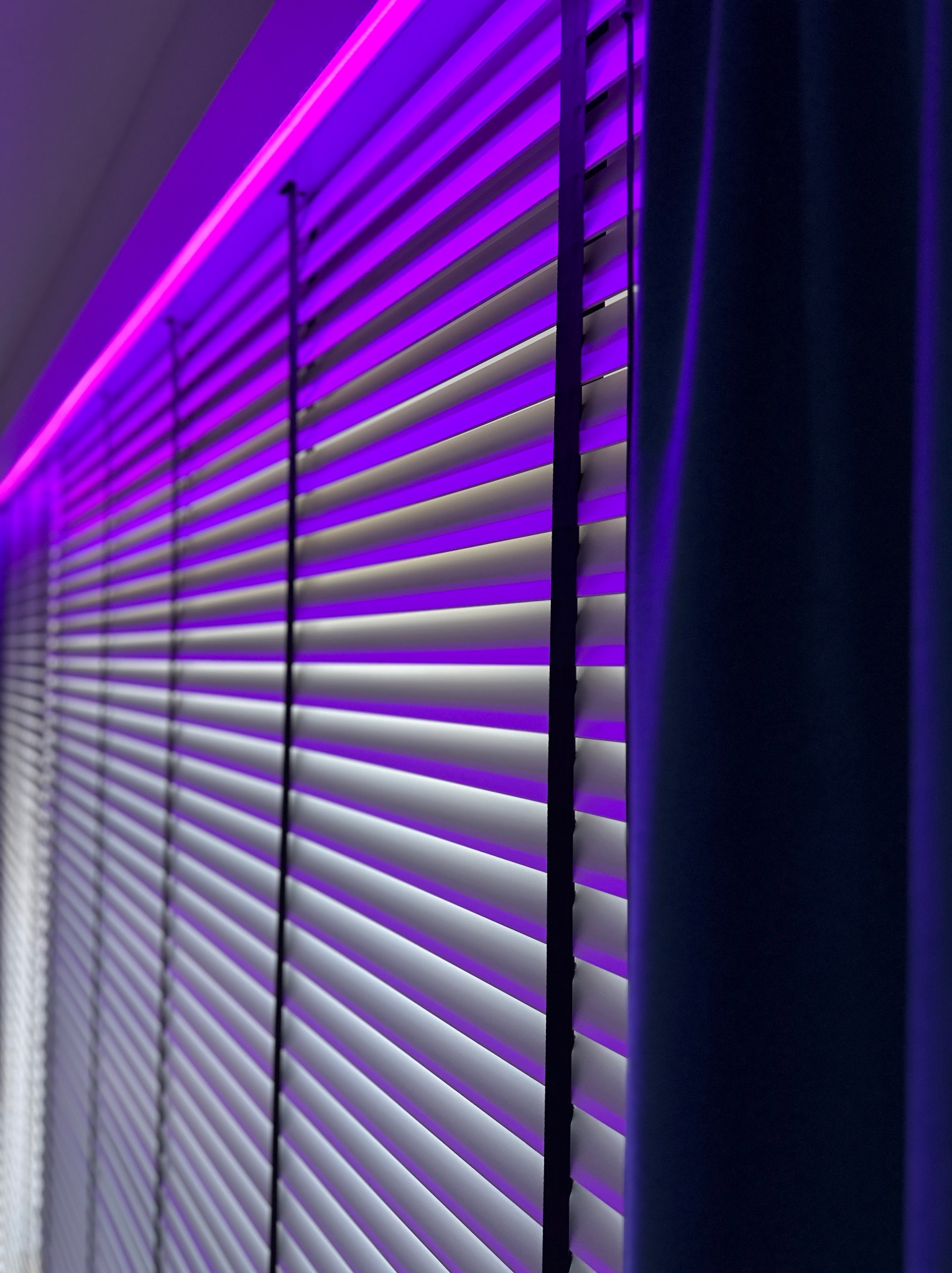 Białe poziome lamele okienne podświetlone intensywnym fioletowym światłem LED, z fragmentem zasłony po prawej stronie kadru.