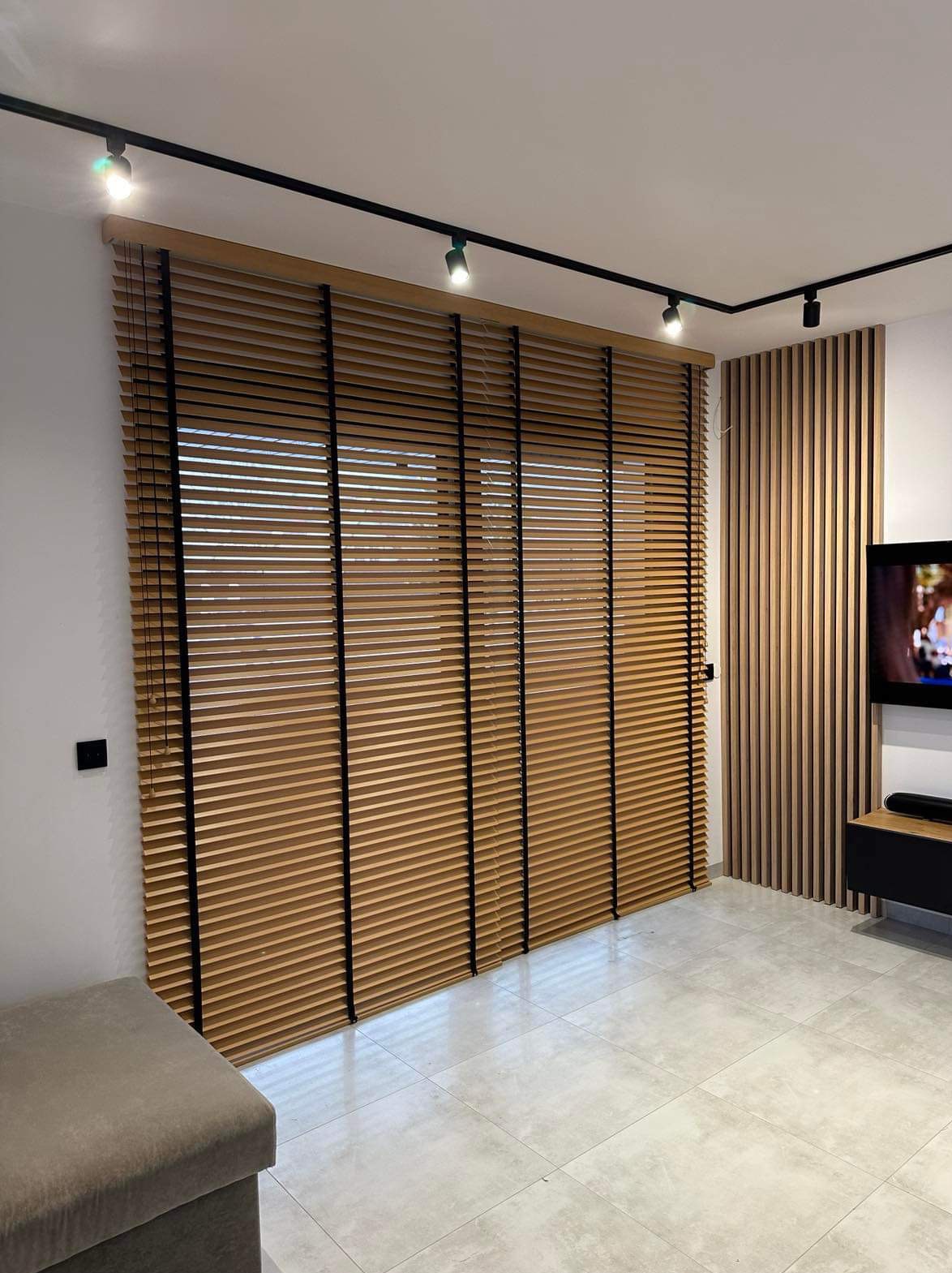 Zamknięte drewniane żaluzje z czarnymi sznurkami w nowoczesnym salonie z drewnianą ścianą dekoracyjną i oświetleniem szynowym.