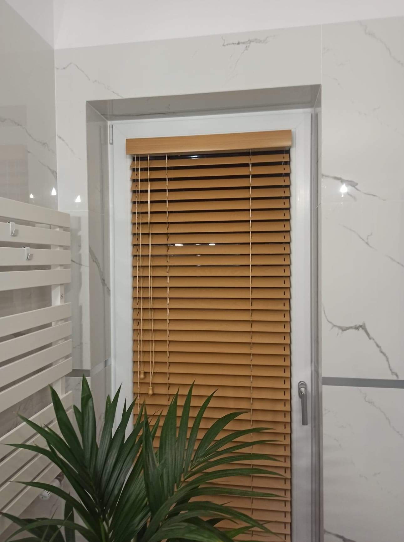 Jasnobrązowe listewki w oknie, z białymi ramami i marmurowymi płytkami w tle, oraz zielona roślina w dolnym rogu.