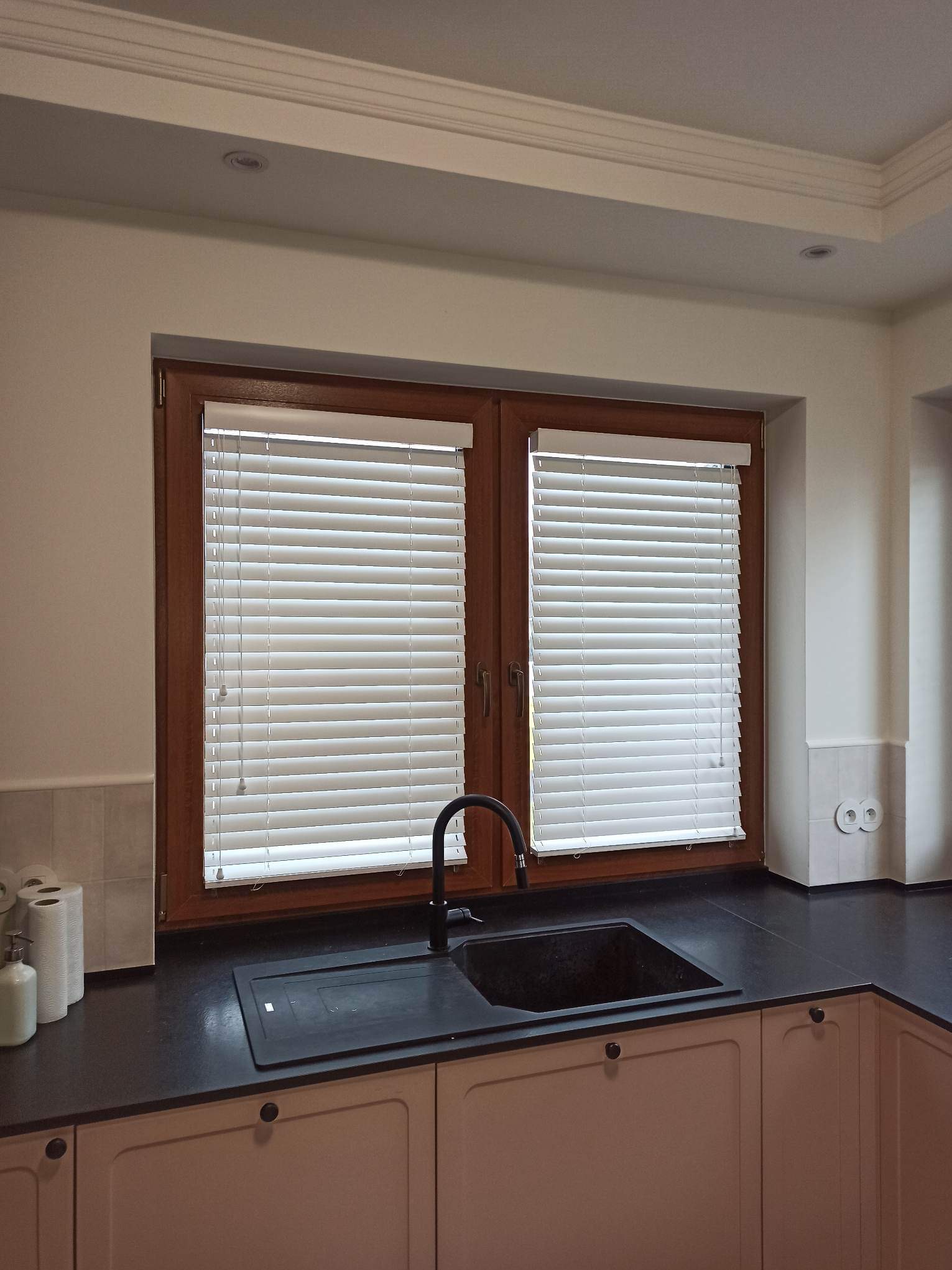 Kuchnia z oknem wyposażonym w poziome, białe listewki regulujące dopływ światła, widoczny zlew i czarna bateria.