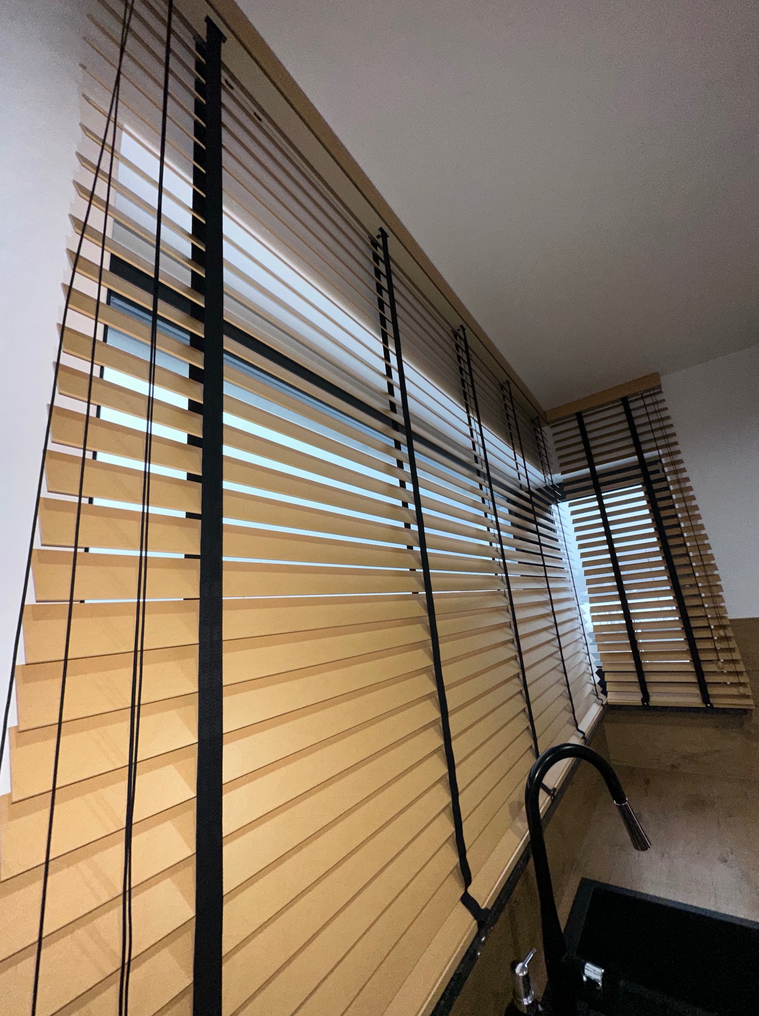 Jasne, drewniane lamele z czarnymi taśmami w oknie, widok z dołu, fragment zlewu na pierwszym planie.