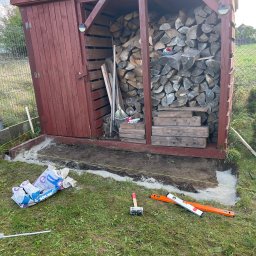 Oskar Żemojdzin - Przygotowanie podłoża pod budowę, widoczne worki z zaprawą SOLBET, poziomica, gumowy młotek, sznurek wyznaczający granice, częściowo wylany beton, drewniana wiata na drewno w tle.