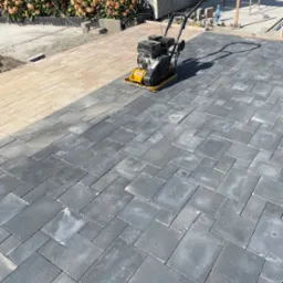 Układanie ciemnoszarej kostki brukowej w regularnym wzorze, zagęszczarka wibracyjna na powierzchni, widoczny fragment jasnej kostki i rabata kwiatowa w tle.