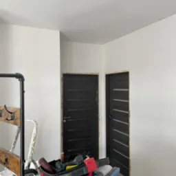 Białe, świeżo wyszpachlowane ściany w pokoju z dwoma ciemnymi drzwiami. Na pierwszym planie widoczne narzędzia i akcesoria malarskie, w tym drabina i kuweta.