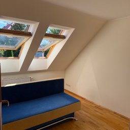 Oskar Żemojdzin - Jasne poddasze z drewnianą podłogą i niebieską sofą. Dwa okna dachowe wpuszczają światło. Świeżo pomalowane ściany w odcieniach beżu. Widoczne listwy przypodłogowe.