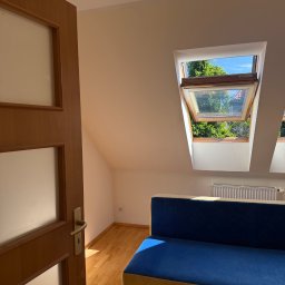 Oskar Żemojdzin - Jasne poddasze z drewnianą podłogą i otwartym oknem dachowym, przez które widać zieleń. Częściowo widoczna niebieska sofa i drewniane drzwi.