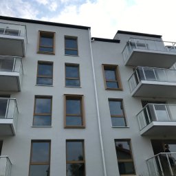 Oskar Żemojdzin - Elewacja budynku wielorodzinnego z balkonami o szklanych balustradach i oknami w drewnianych ramach. Widok z dołu na jasną fasadę i błękitne niebo.