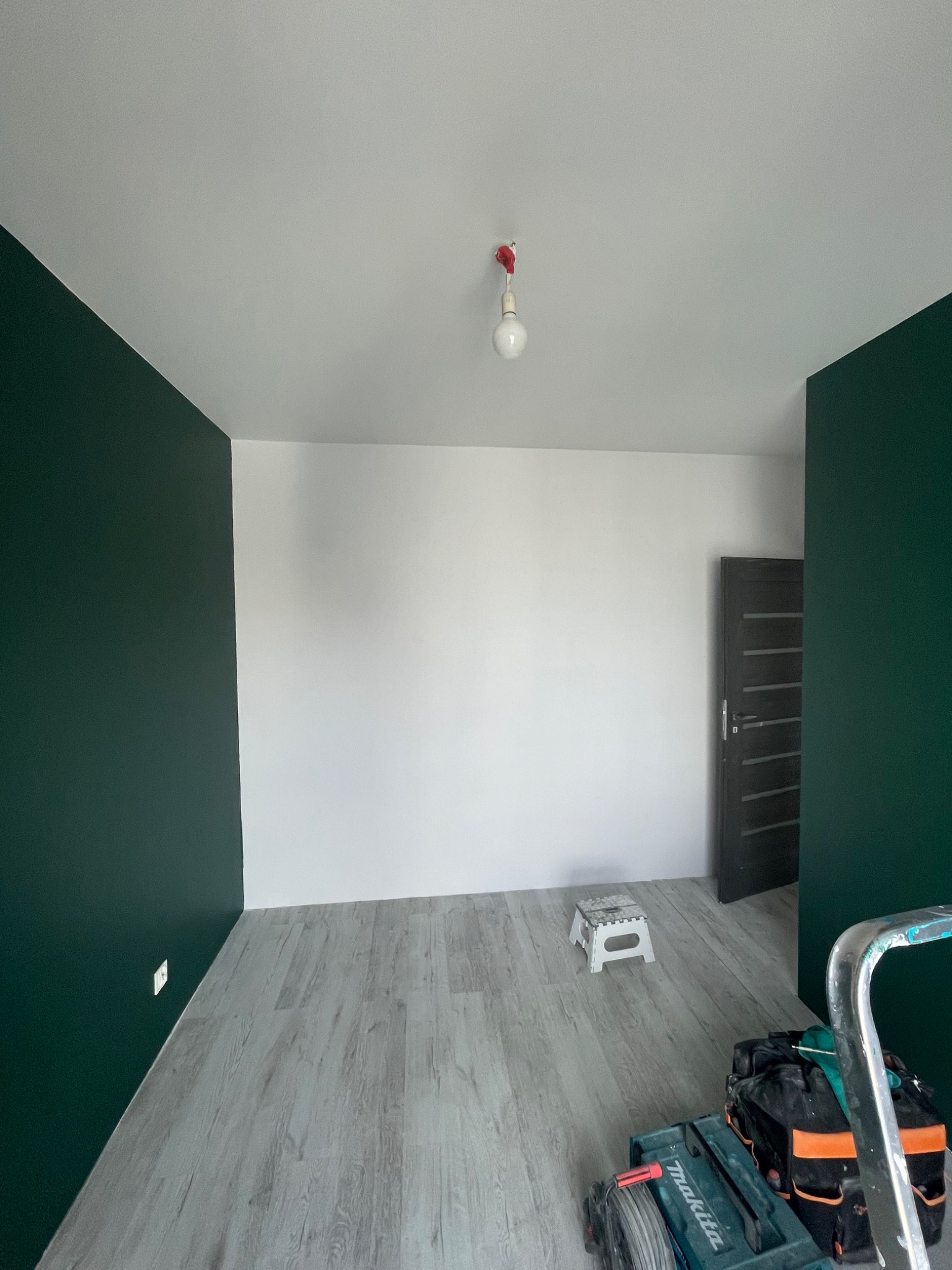 Pokój po częściowym pomalowaniu ścian na ciemnozielony kolor, widoczna drabina, narzędzia i stołek, podłoga wyłożona panelami imitującymi drewno.