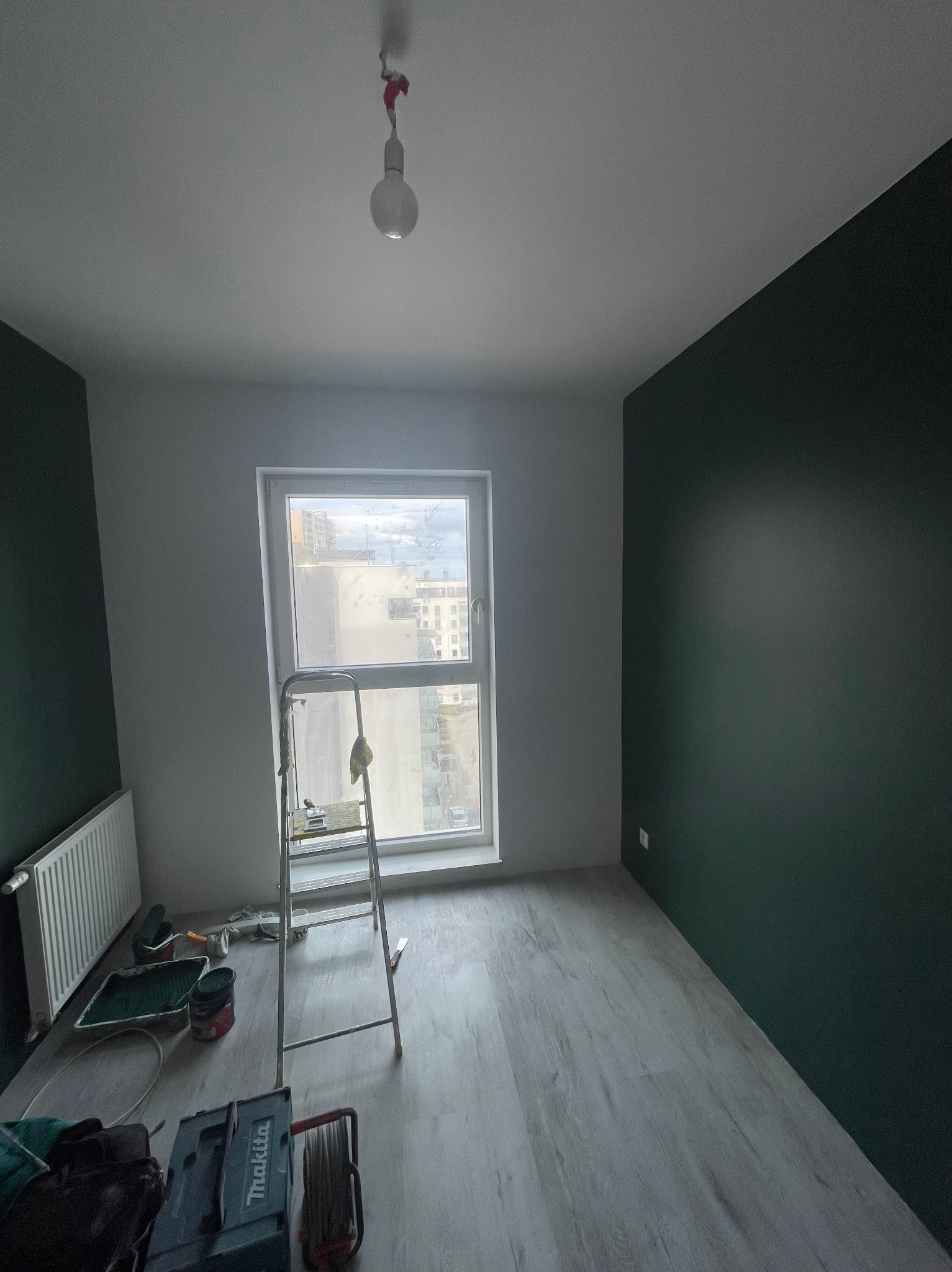 Pokój po częściowym pomalowaniu ścian w Dzierzgoniu: jedna ściana biała, druga w odcieniach zieleni, drabina przy oknie, narzędzia malarskie w rogu.