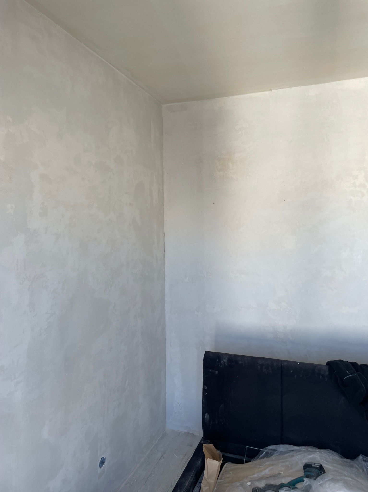 Świeżo wyszpachlowana ściana w pokoju z widocznymi śladami szpachli, przygotowana do dalszych prac wykończeniowych, częściowo zasłonięta przez zagłówek łóżka i narzędzia malarskie.