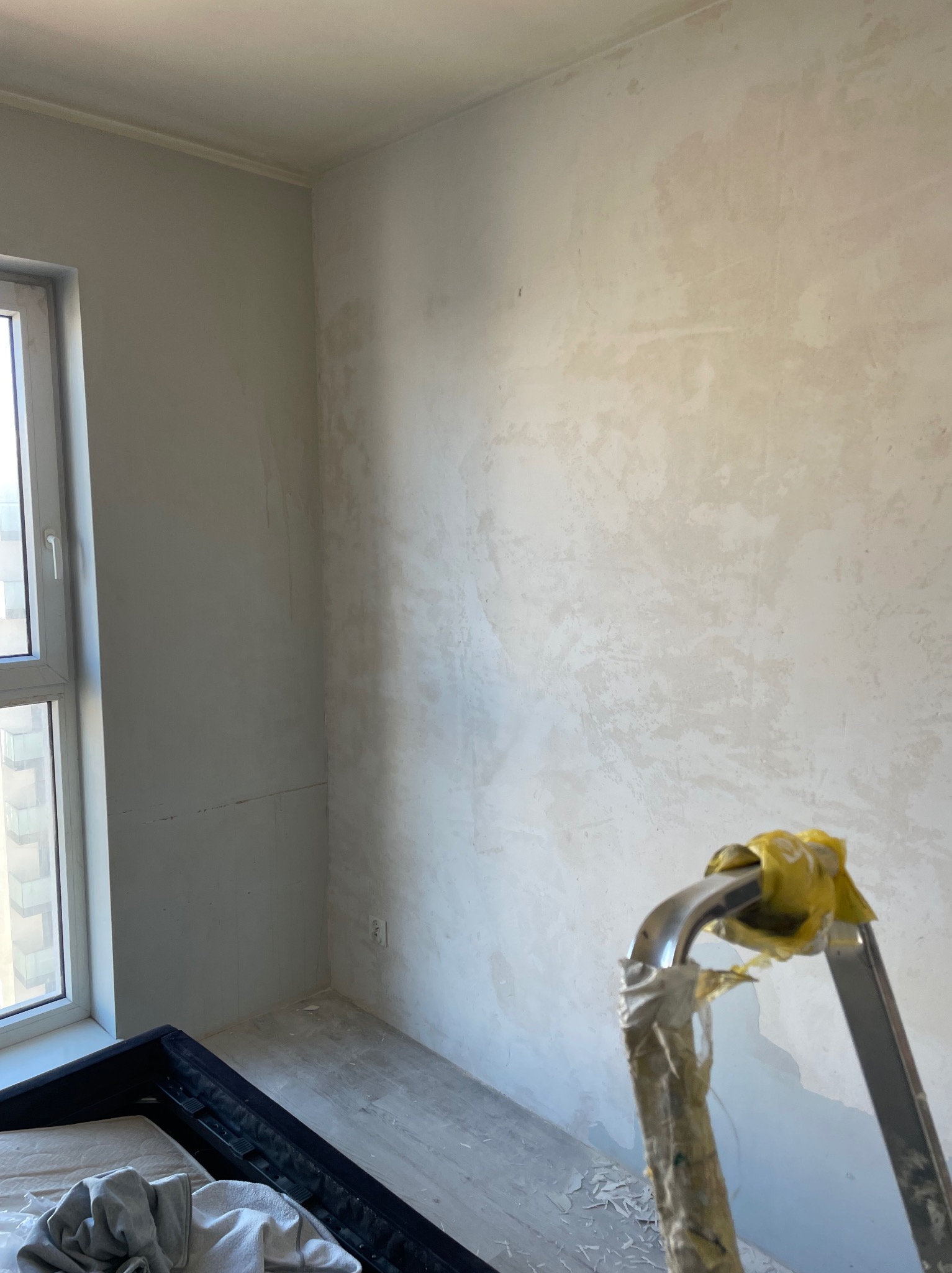 Pokój w trakcie remontu: ściany po nałożeniu szpachli, przygotowane do malowania, widoczna drabina malarska z zabezpieczeniem i resztkami materiałów budowlanych, okno z widokiem na budynek, podłoga...