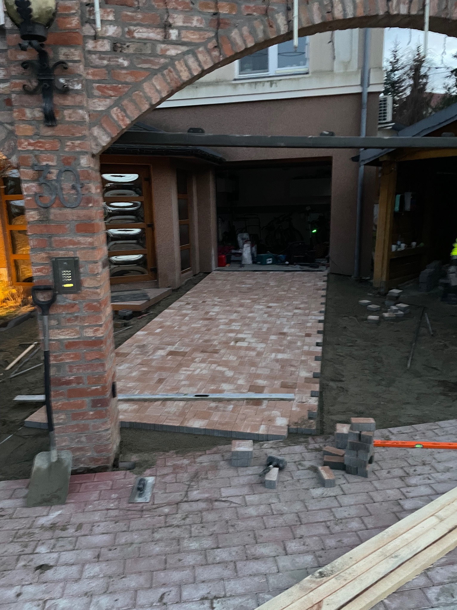 Ceglana ścieżka z kostki brukowej w trakcie układania, widoczna łopata oparta o ceglany filar z numerem domu, poziomica i stosy kostki brukowej w tle, garaż.