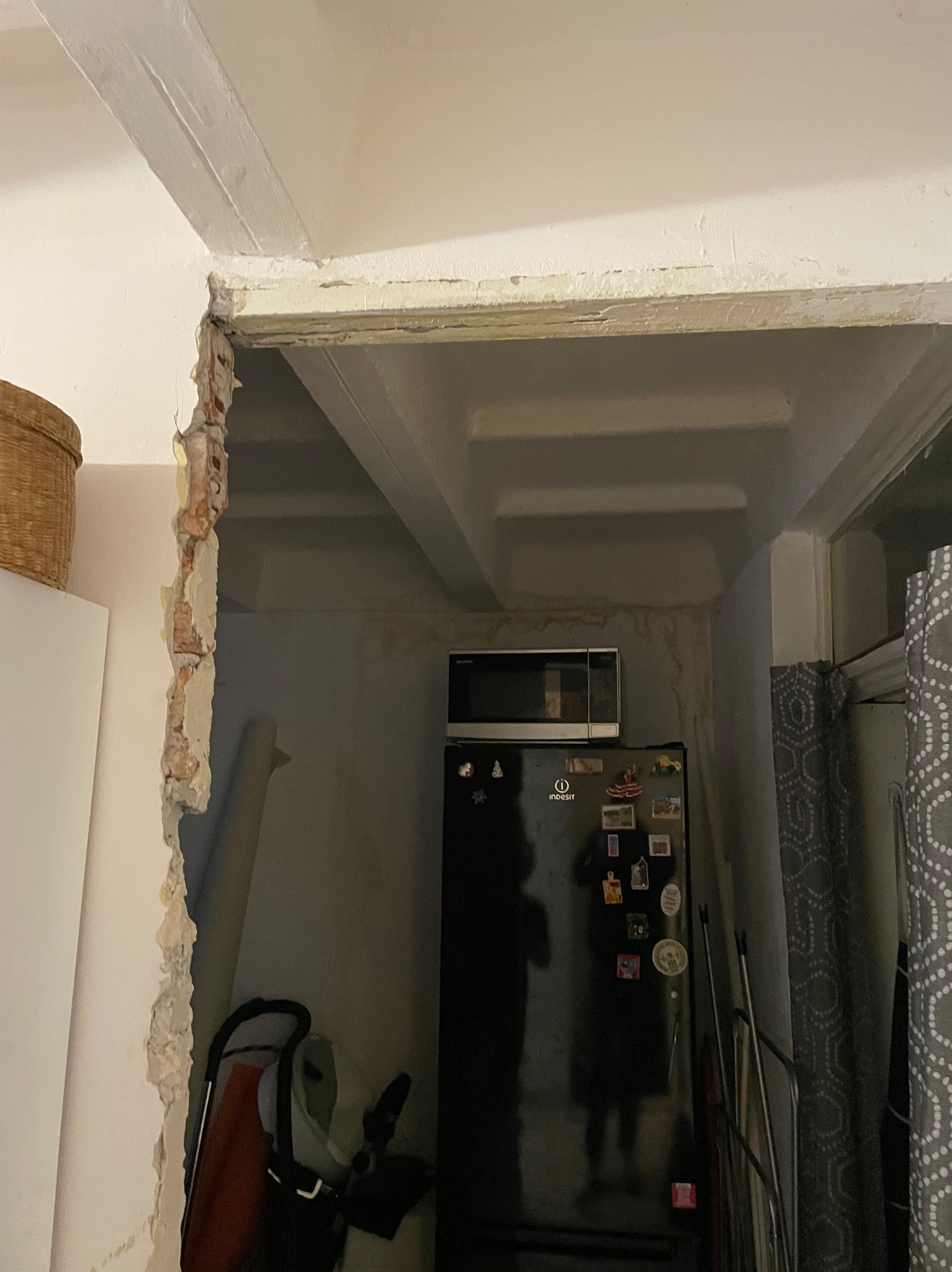 Otwór w ścianie z odsłoniętą cegłą i resztkami tynku, widok na wnętrze z lodówką i mikrofalówką, przygotowanie do remontu.