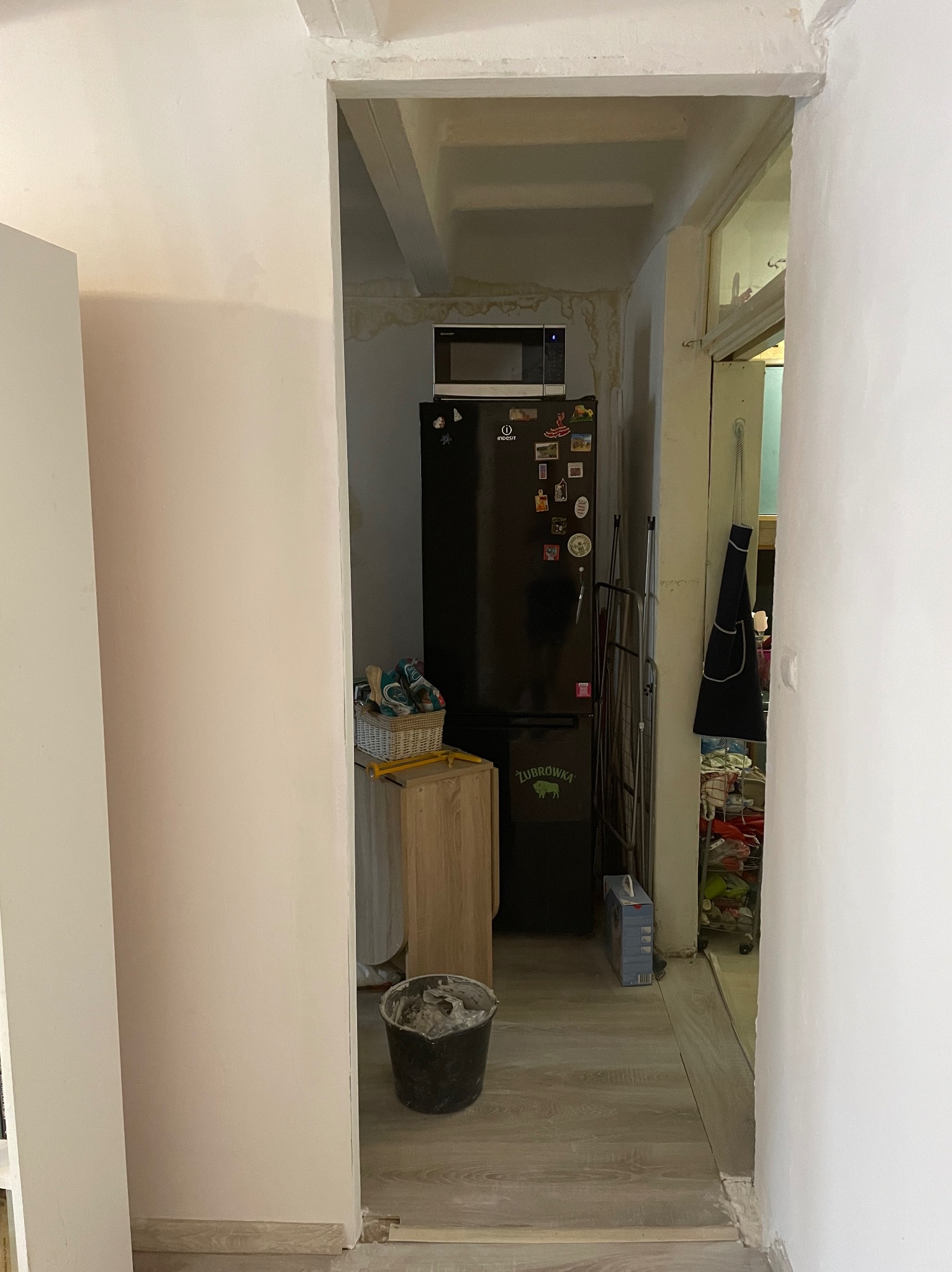 Fragment pokoju z pomalowaną na jasny róż ścianą, widoczny czarny kubeł z resztkami farby, w tle lodówka z magnesami i kuchenka mikrofalowa, ślady wilgoci na suficie.