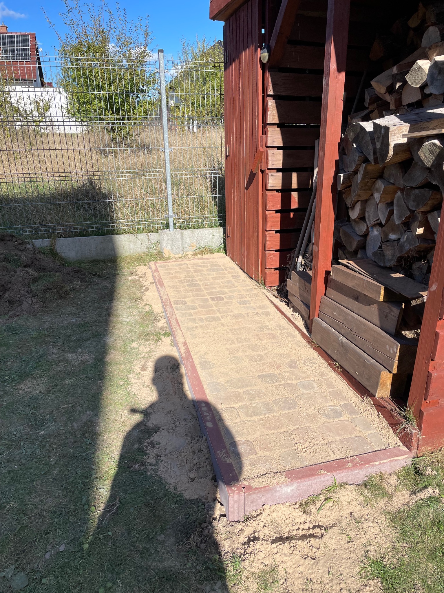 Ułożona ścieżka z kostki brukowej prowadząca do drewnianej drewutni, częściowo zasypana piaskiem, cień osoby w lewym dolnym rogu.
