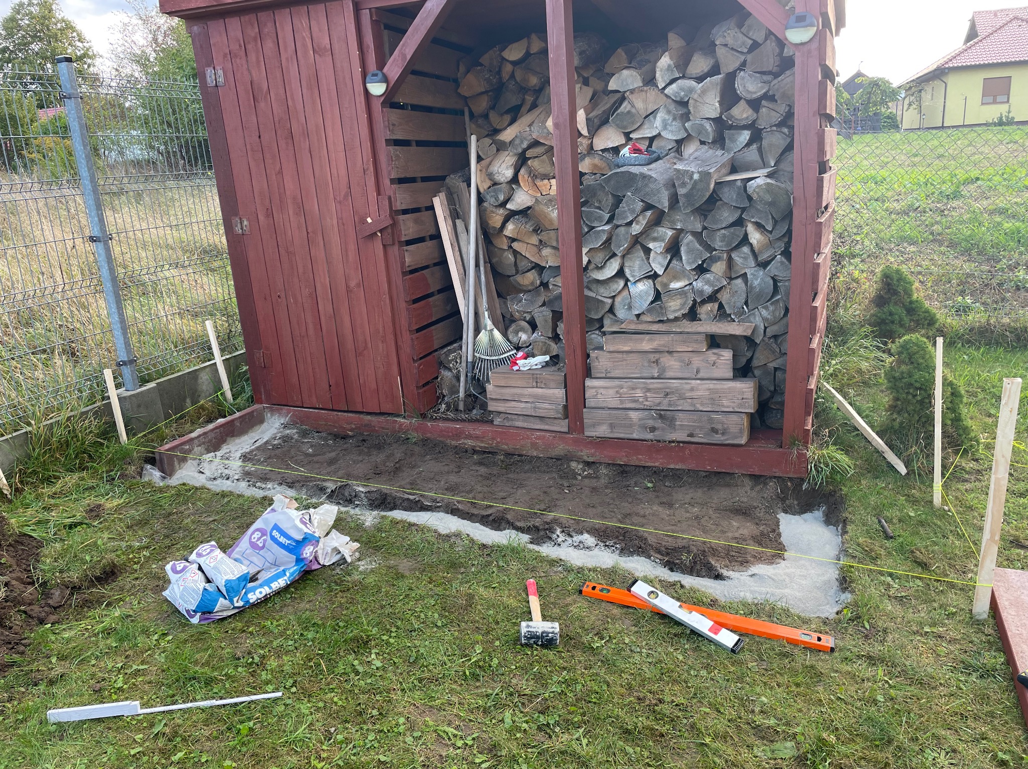 Przygotowanie podłoża pod budowę, widoczne worki z zaprawą SOLBET, poziomica, gumowy młotek, sznurek wyznaczający granice, częściowo wylany beton, drewniana wiata na drewno w tle.