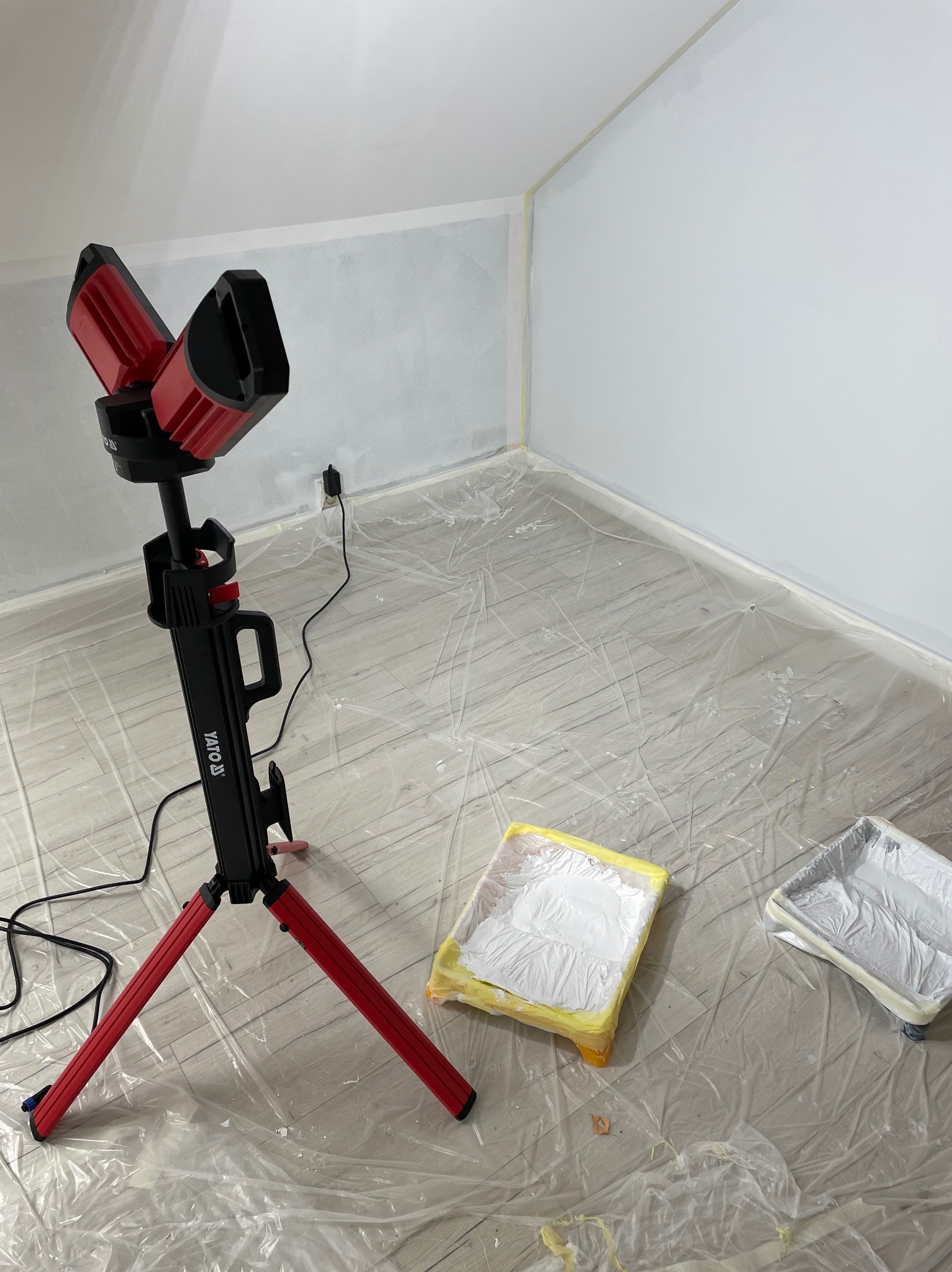 Pokój w trakcie malowania na biało, widoczny czerwony statyw z lampami YATO, kuweta malarska z farbą i folia ochronna na podłodze.