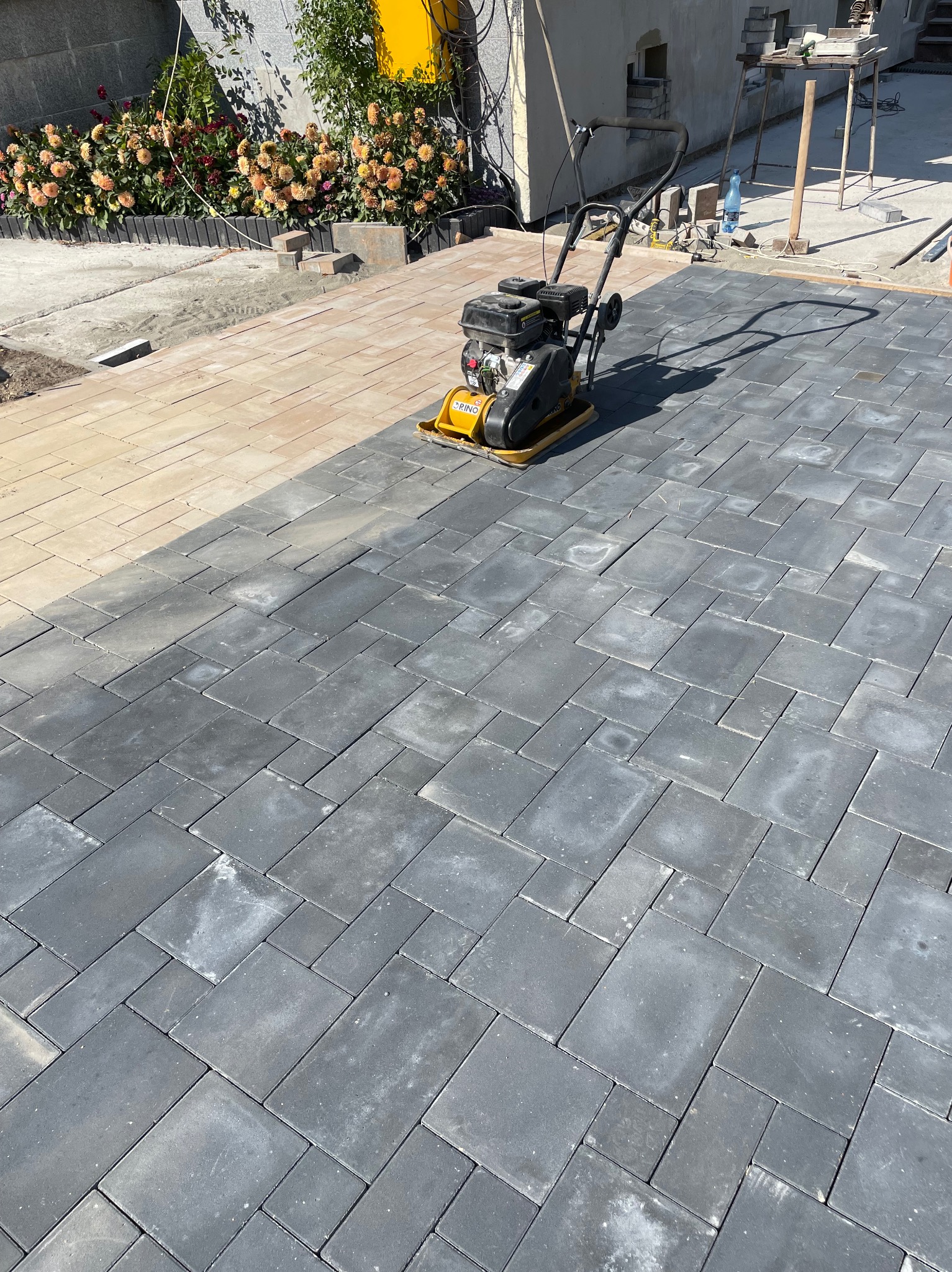 Układanie ciemnoszarej kostki brukowej w regularnym wzorze, zagęszczarka wibracyjna na powierzchni, widoczny fragment jasnej kostki i rabata kwiatowa w tle.