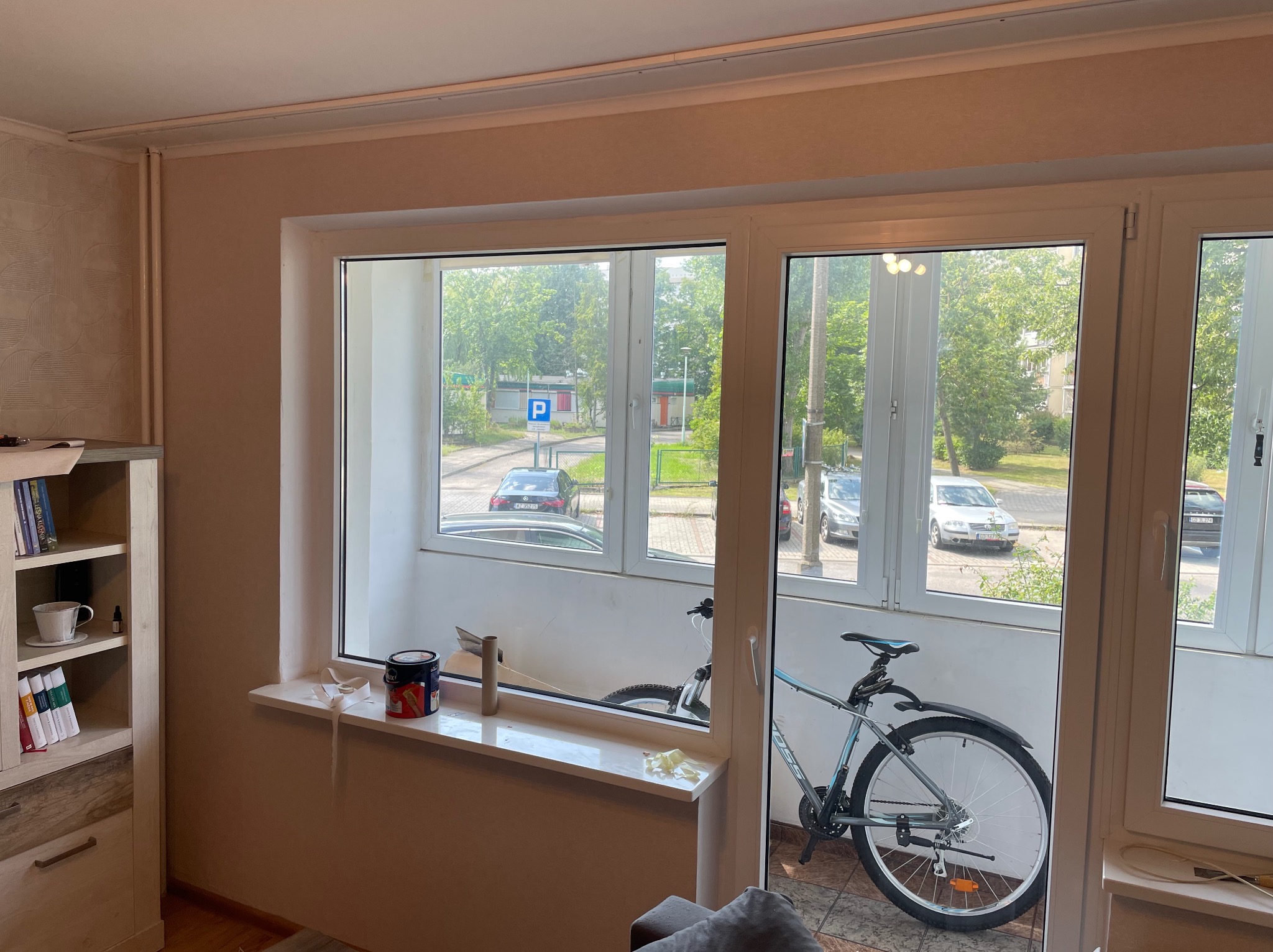 Pokój w trakcie tapetowania: widoczna puszka farby na parapecie, ślady po taśmie malarskiej i rower na balkonie. W tle zabudowa i okno z widokiem na ulicę.