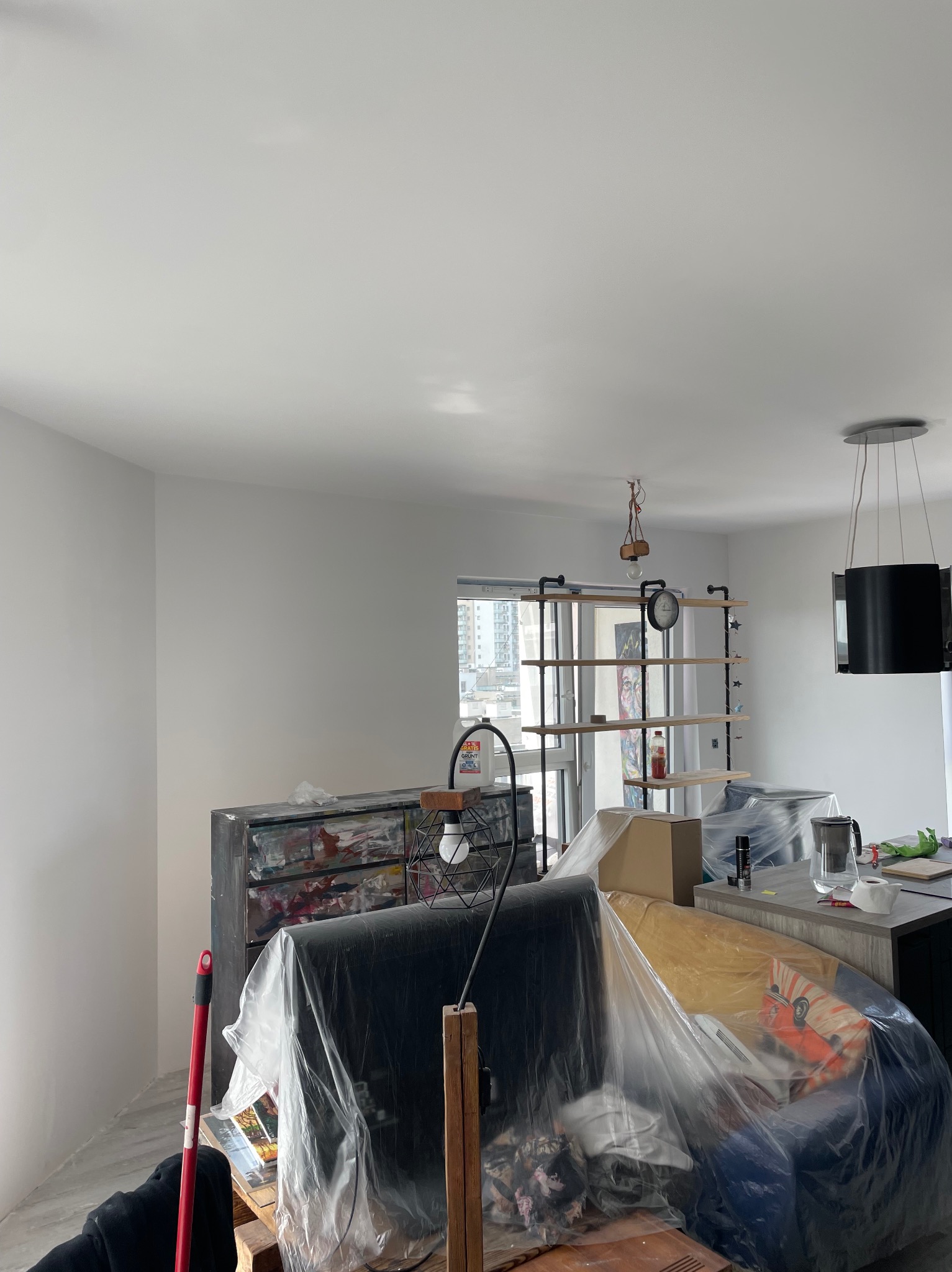 Wnętrze po szpachlowaniu: gładkie, białe ściany i sufit. Meble zabezpieczone folią malarską. Widoczny regał i lampa w stylu industrialnym. Świeżo po remoncie.