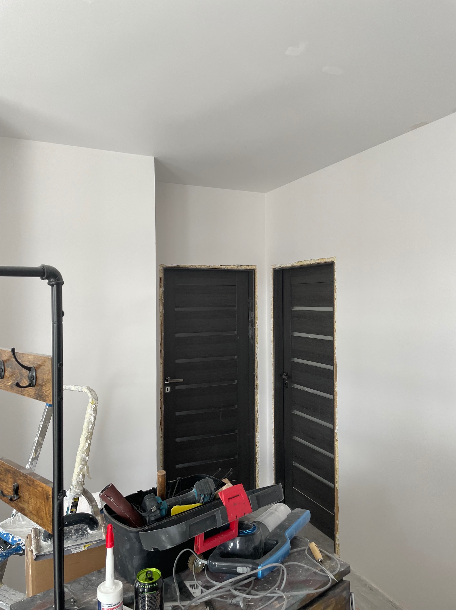 Białe, świeżo wyszpachlowane ściany w pokoju z dwoma ciemnymi drzwiami. Na pierwszym planie widoczne narzędzia i akcesoria malarskie, w tym drabina i kuweta.
