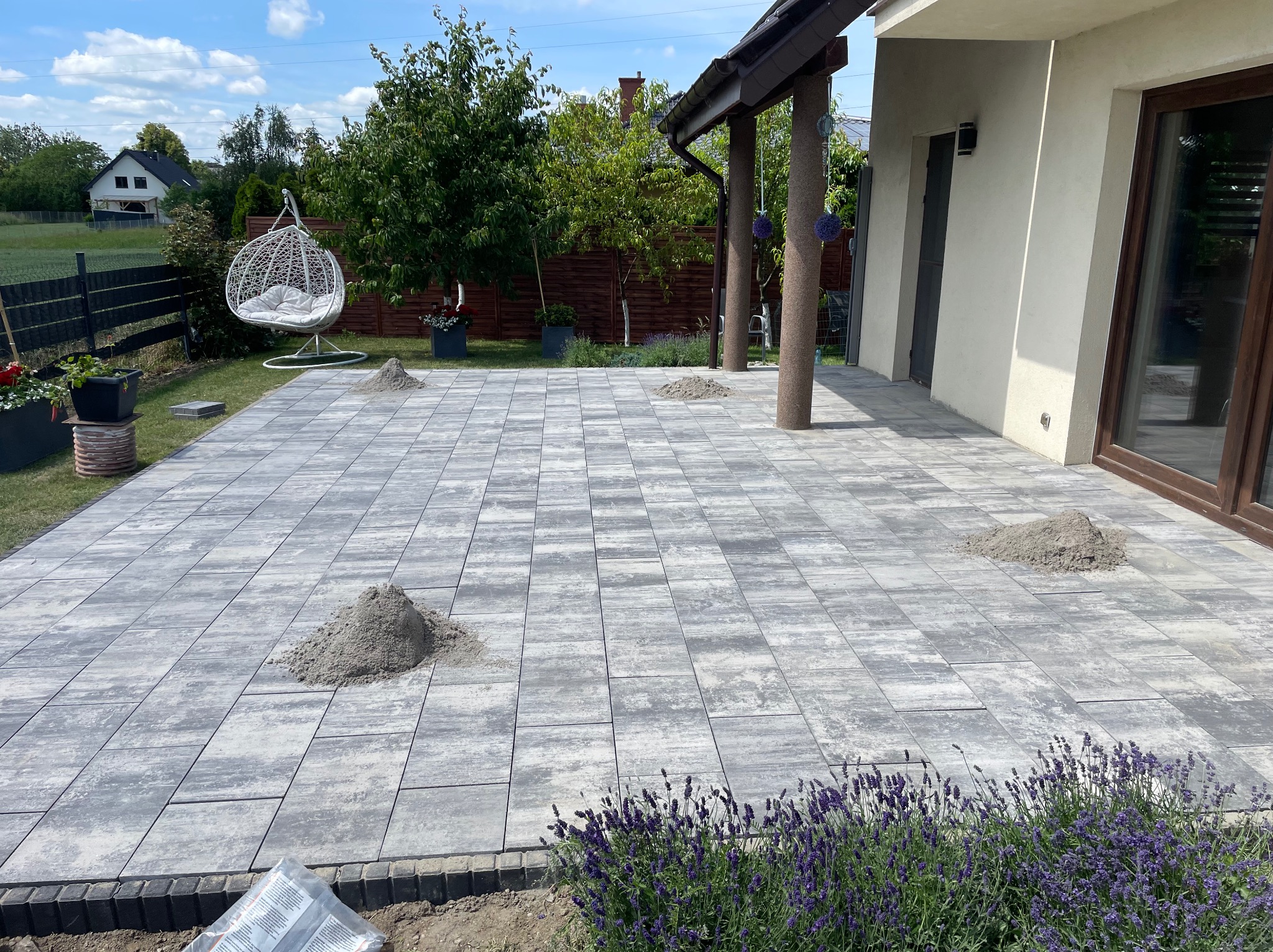 Ułożona szara kostka brukowa na posesji, z widocznymi kopcami piasku do wypełnienia szczelin. W tle dom z elementami architektury ogrodowej i wiszące krzesło.
