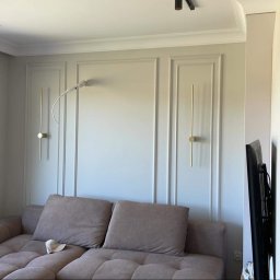 USŁUGI REMONTOWO-WYKOŃCZENIOWE ART.-MAT DAWID MATYKIEWICZ - Ściana w salonie z dekoracyjnymi panelami i złotymi lampami ściennymi, widoczny fragment sofy i okno z widokiem na zewnątrz, kabel lampy wystający ze ściany przed ostatecznym wykończeniem.