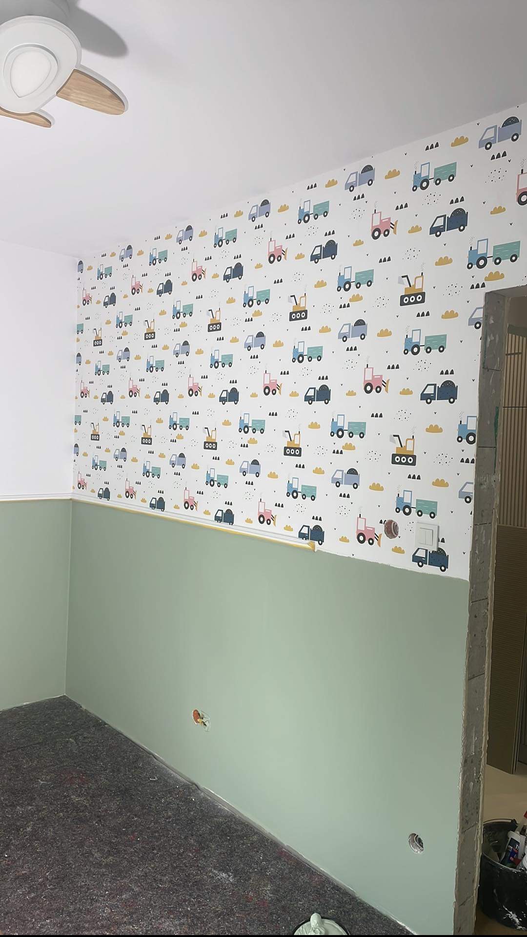 Ściana w pokoju dziecięcym z dolną częścią pomalowaną na zielono i górną wytapetowaną w kolorowe pojazdy budowlane na białym tle, widoczny fragment sufitu z lampą.