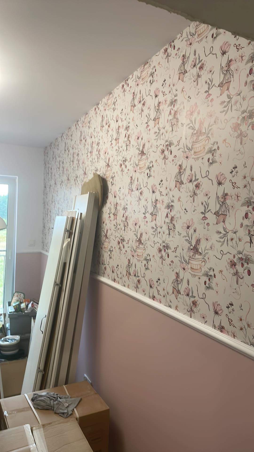Pokój po remoncie z tapetą w kwiaty i myszy, dolna część ściany pomalowana na różowo, przy ścianie oparte białe drzwi.
