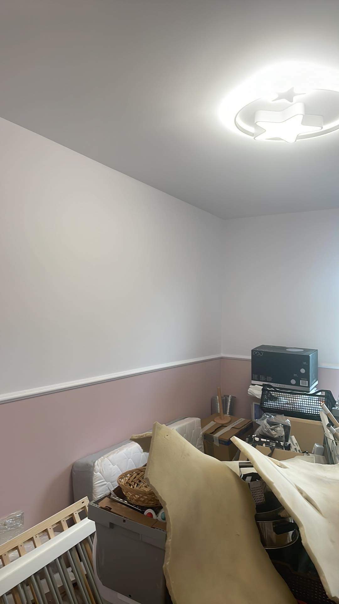 Pokój w trakcie remontu: ściany pomalowane na biało i różowo, widoczna listwa przypodłogowa, na podłodze bałagan, w tym fragmenty pianki, materac, koszyk wiklinowy i elementy łóżeczka dziecięcego.
