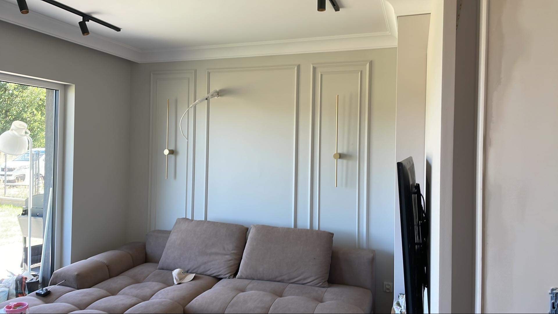 Ściana w salonie z dekoracyjnymi panelami i złotymi lampami ściennymi, widoczny fragment sofy i okno z widokiem na zewnątrz, kabel lampy wystający ze ściany przed ostatecznym wykończeniem.