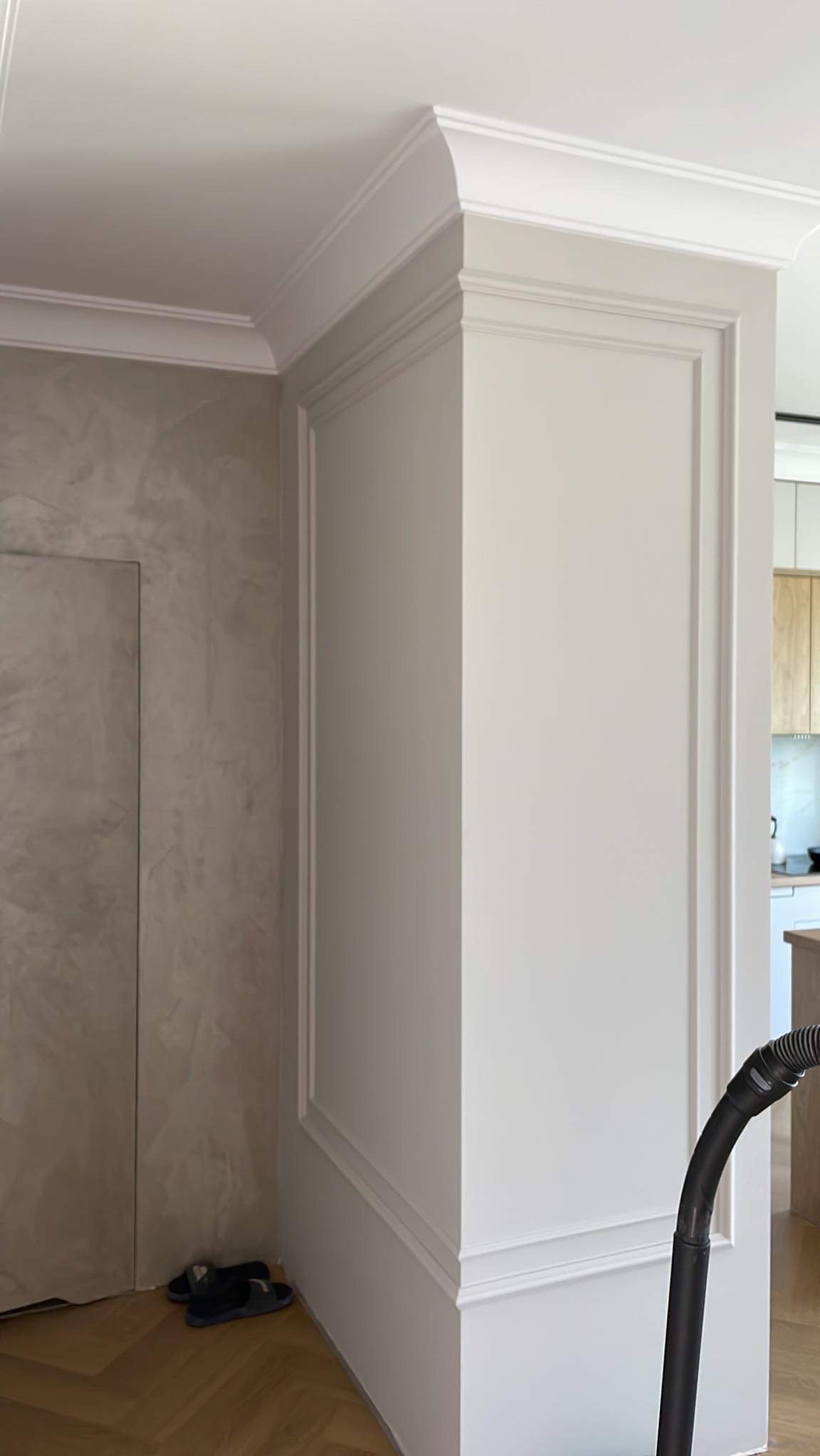 Wykończony narożnik pokoju z dekoracyjną sztukaterią, ściana z efektem betonu, parkiet w jodełkę i fragment odkurzacza w tle.