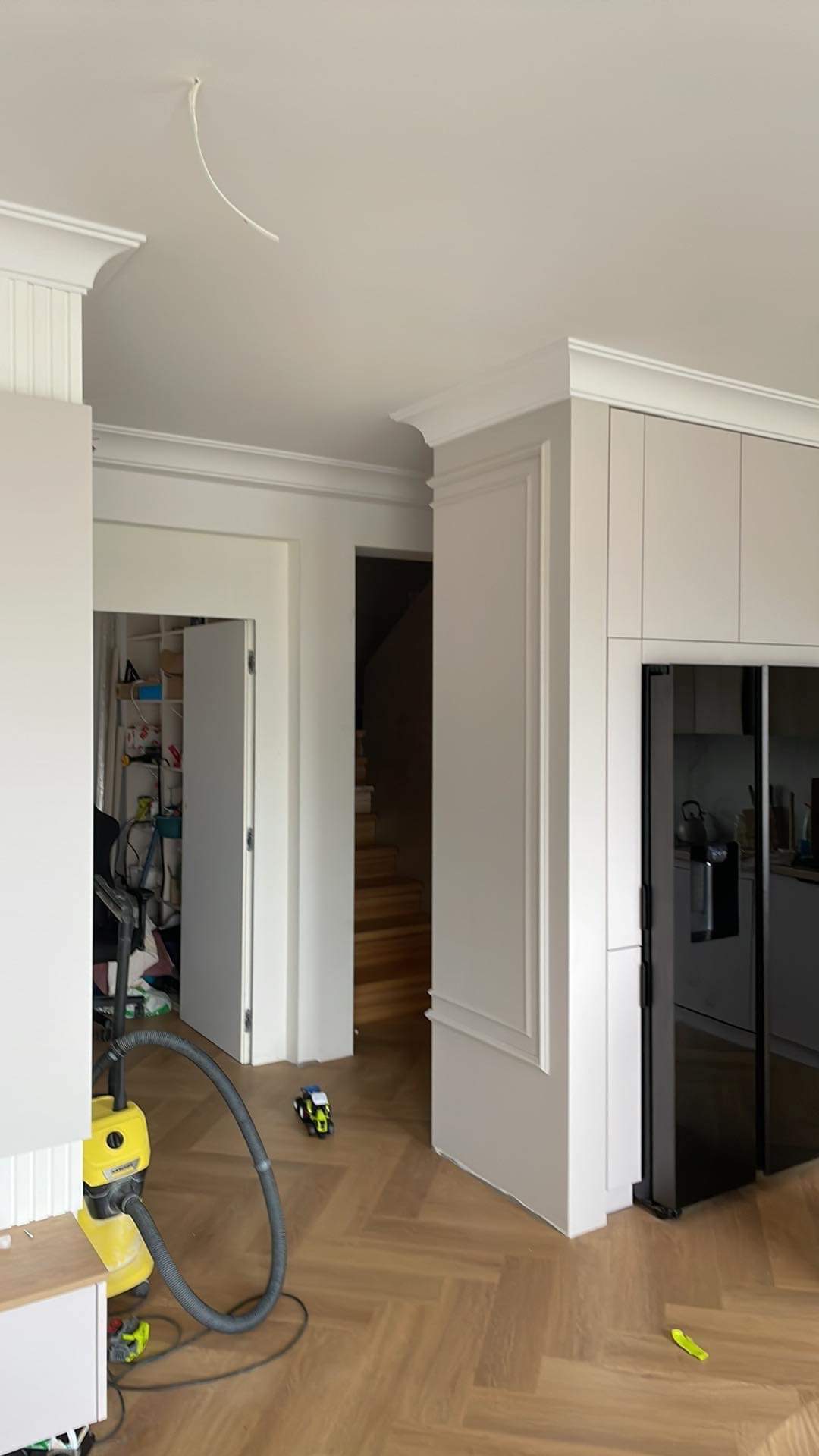 Wnętrze w trakcie wykończenia: widoczna podłoga ułożona w jodełkę, białe listwy przypodłogowe, drzwi wewnętrzne z ozdobnymi ramami, częściowo zabudowana kuchnia z czarną lodówką i żółty odkurzacz...