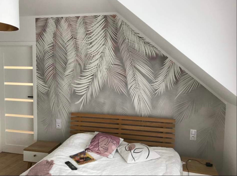Sypialnia na poddaszu z tapetą w motywie palmowych liści, drewnianym zagłówkiem łóżka i minimalistycznymi poduszkami z motywem twarzy.