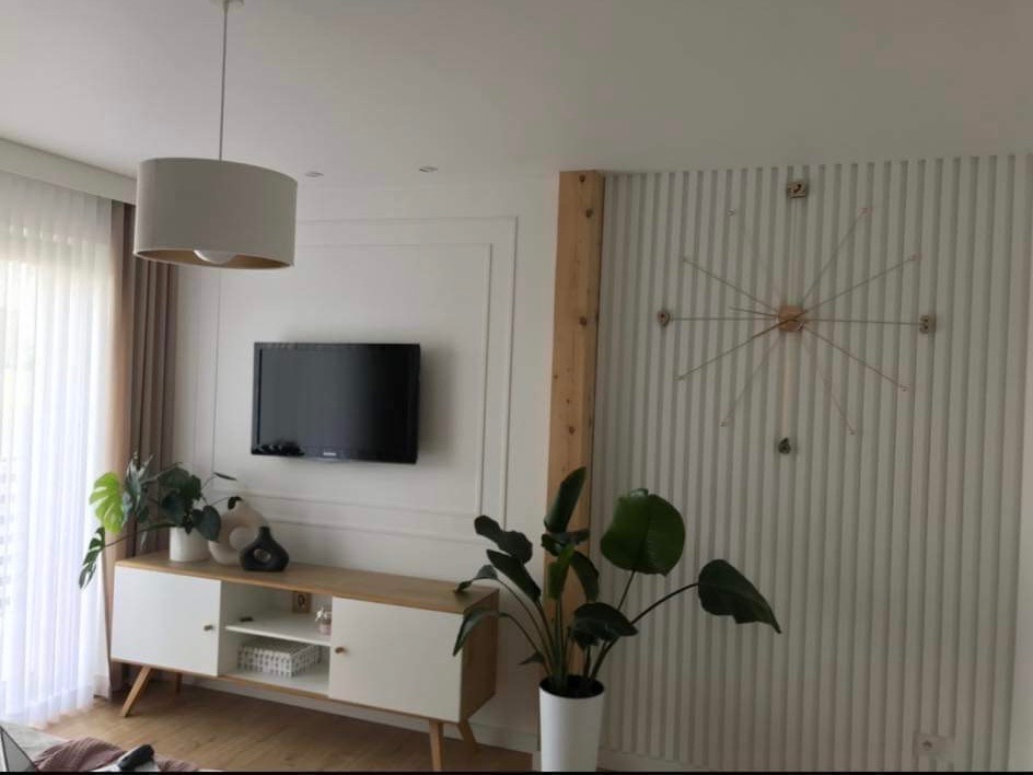 Nowoczesny salon z białymi lamelami na ścianie, minimalistycznym zegarem w kształcie słońca, wiszącą lampą z abażurem, szafką RTV w stylu skandynawskim i roślinami doniczkowymi.