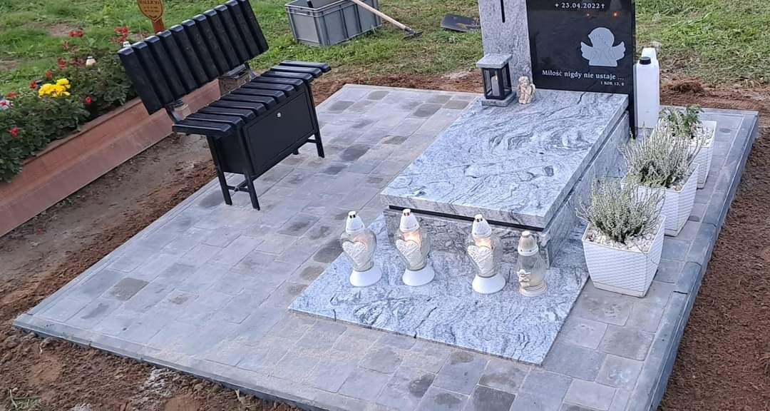 Wykończony nagrobek z szarego granitu, otoczony świeżo ułożoną kostką brukową w odcieniach szarości, z czarną ławką z metalowymi elementami po lewej stronie i czterema lampionami w kształcie serc...