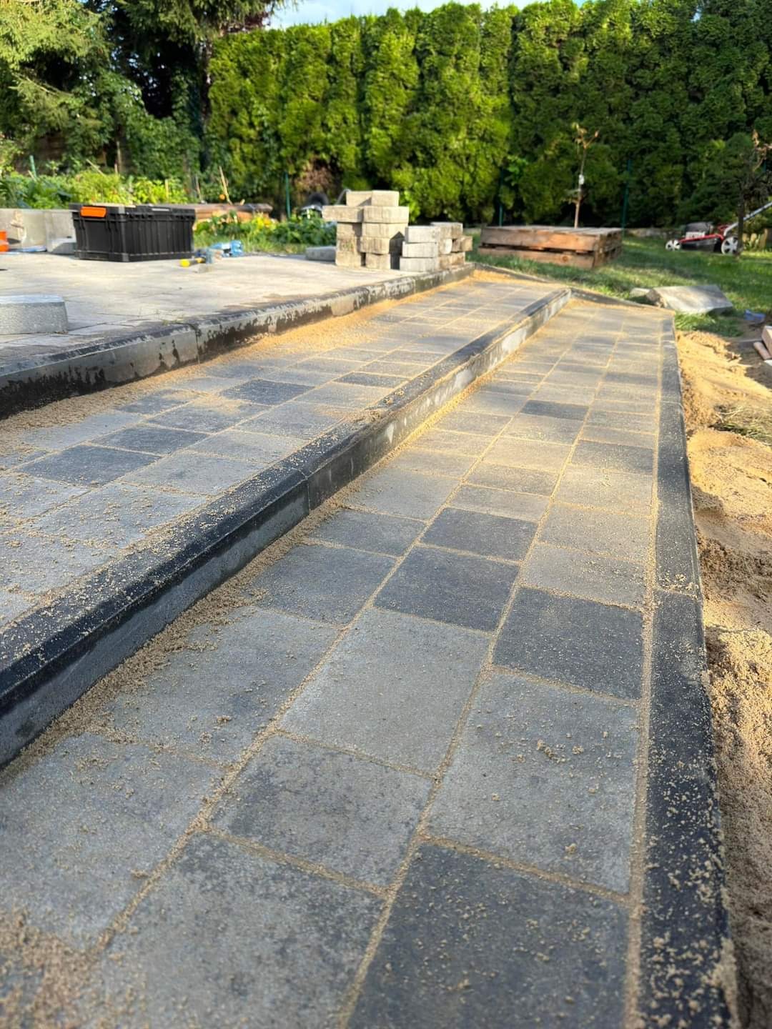 Świeżo ułożony chodnik z szarych kostek brukowych z czarnym obrzeżem, zasypany piaskiem, na tle zielonego ogrodu z tujami i stertą kostki.